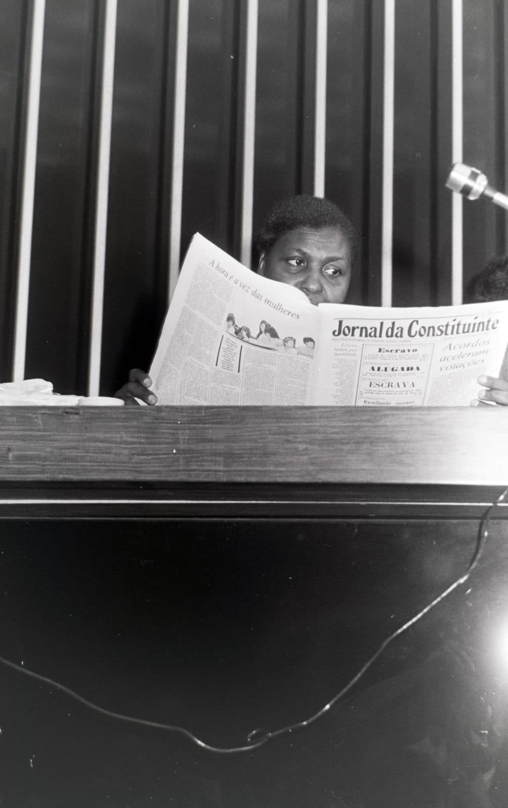 Benedita atrás do “Jornal da Constituinte” aberto, só os olhos aparecem sobre o jornal. Manchete visível: “A hora e a vez das mulheres”. P&B, vertical, altíssima resolução.