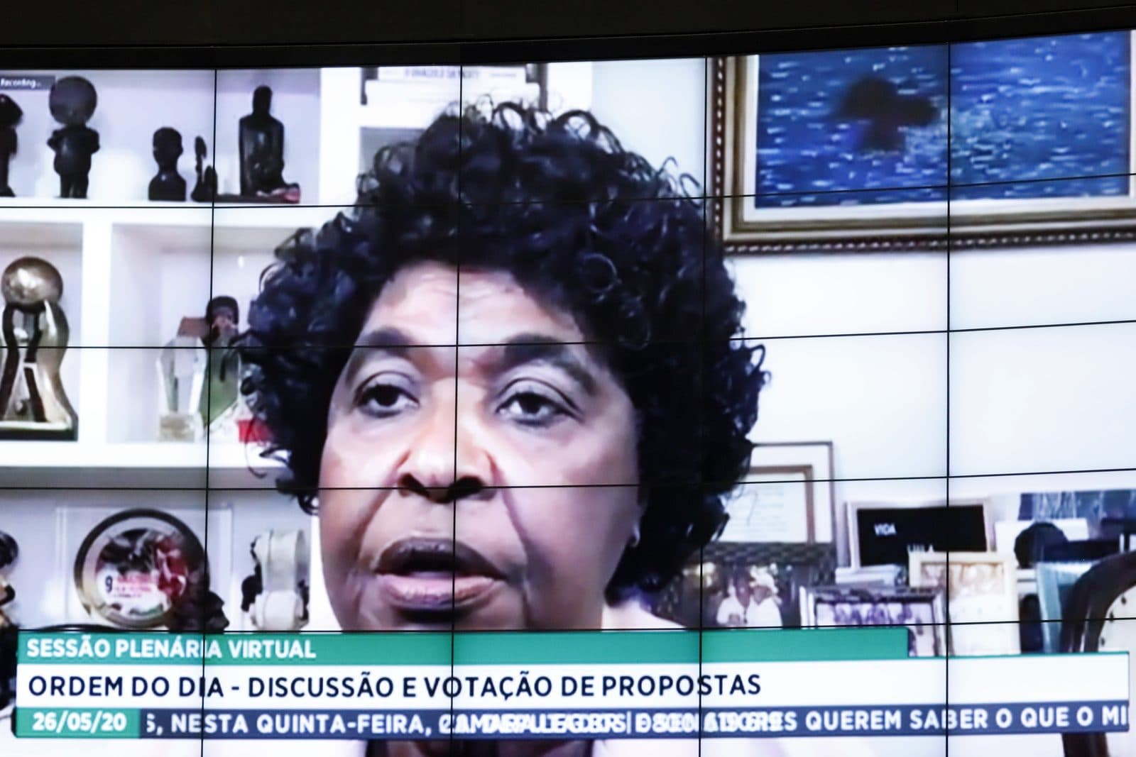 Benedita no telão do plenário durante sessão virtual da pandemia, ao fundo estante com esculturas africanas. Tarja na imagem: “SESSÃO PLENÁRIA VIRTUAL — ORDEM DO DIA — DISCUSSÃO E VOTAÇÃO DE PROPOSTAS — 26/05/20”.