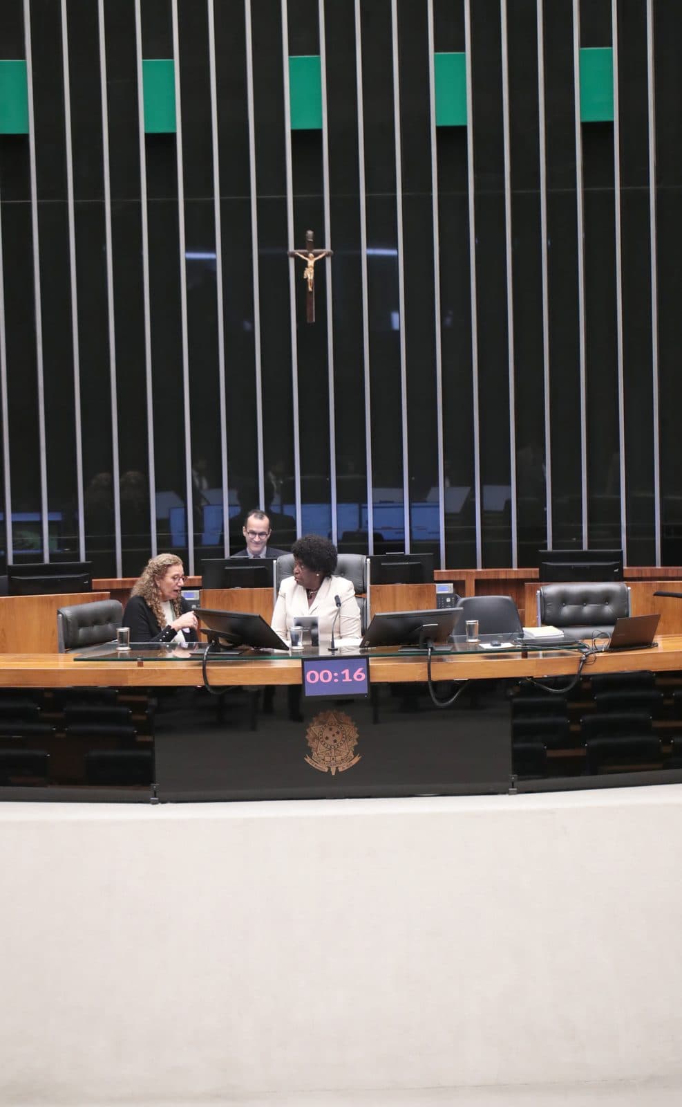 Plano aberto vertical do plenário da Câmara: Benedita sentada à Mesa Diretora, blazer claro, conversando com outra deputada. Plano de ambiente institucional.