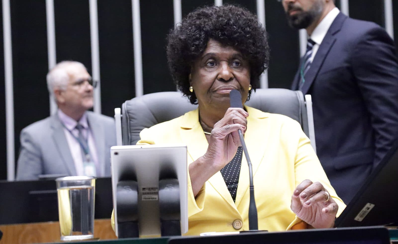 Retrato atual quase frontal: Benedita sentada à mesa do plenário segurando o microfone junto ao rosto, blazer amarelo, olhar firme.